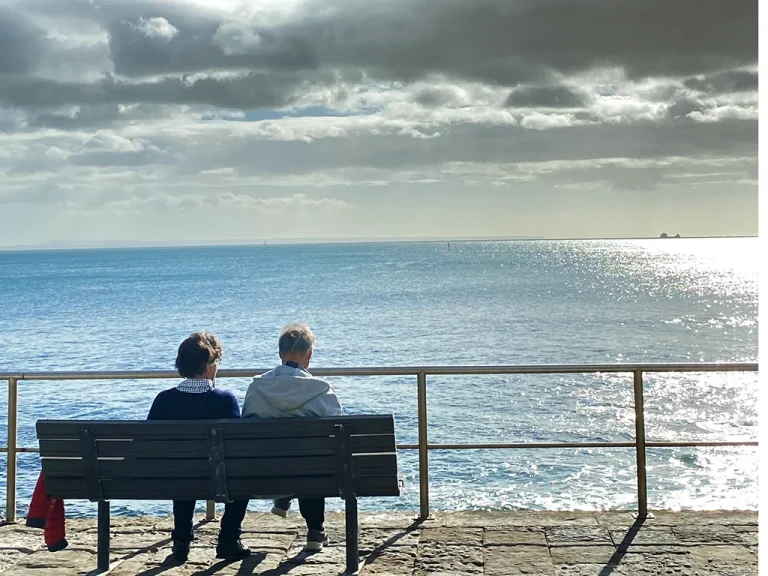couple sitting on a bench looking at the ocean, blue sky, white clouds